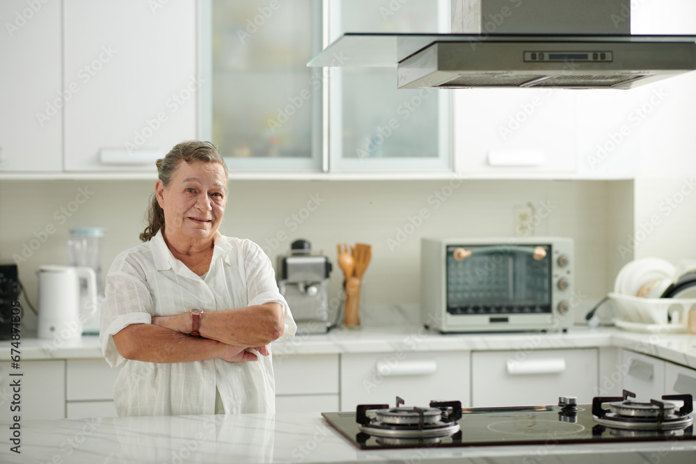 Portrait of smiling elderly woman standing at kitchen counter with arms crossed after cleaning surfaces