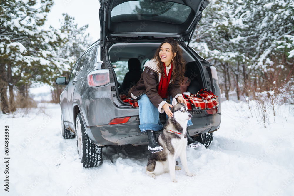 Happy woman with her husky having a picnic in the trunk of a car on a snowy road while traveling by car during the winter holidays. Travel concept.