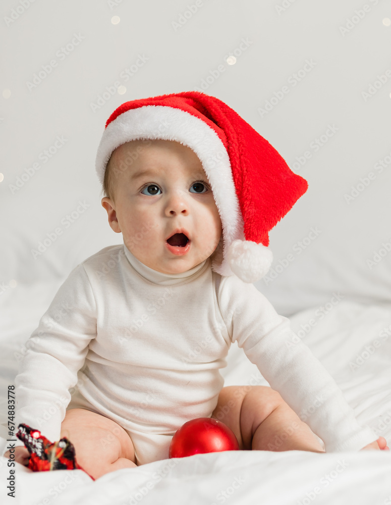 Christmas toddler in Santa hat sitting on bed with gift box and festive baubles Christmas toddler in Santa hat sitting on bed with gift box and festive baubles