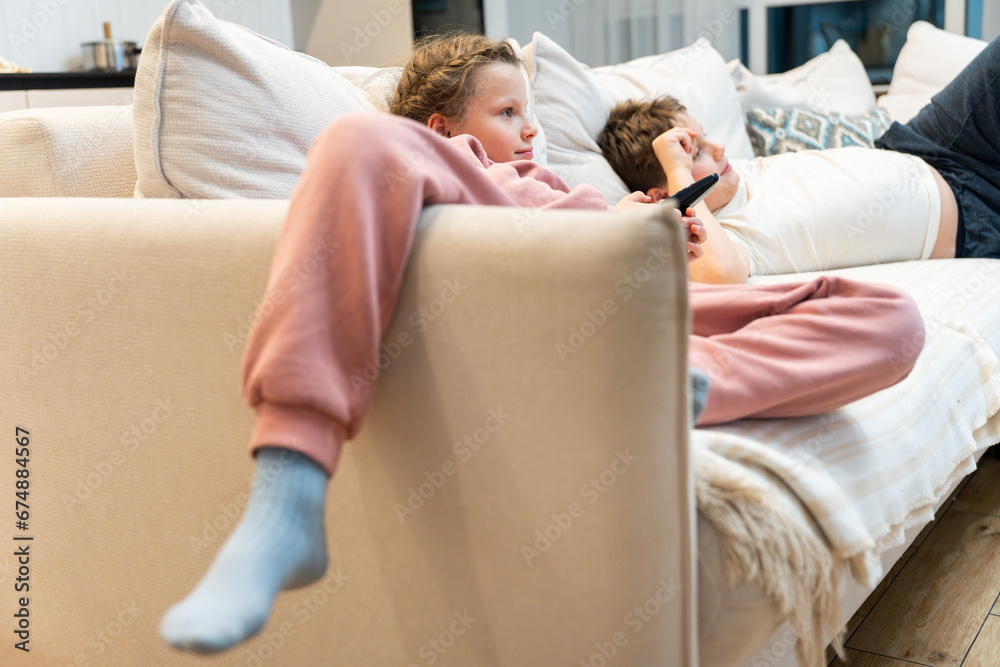 Children lying on the sofa and watching TV interesting cartoon. A girl holding TV remote. Rest in the evening.