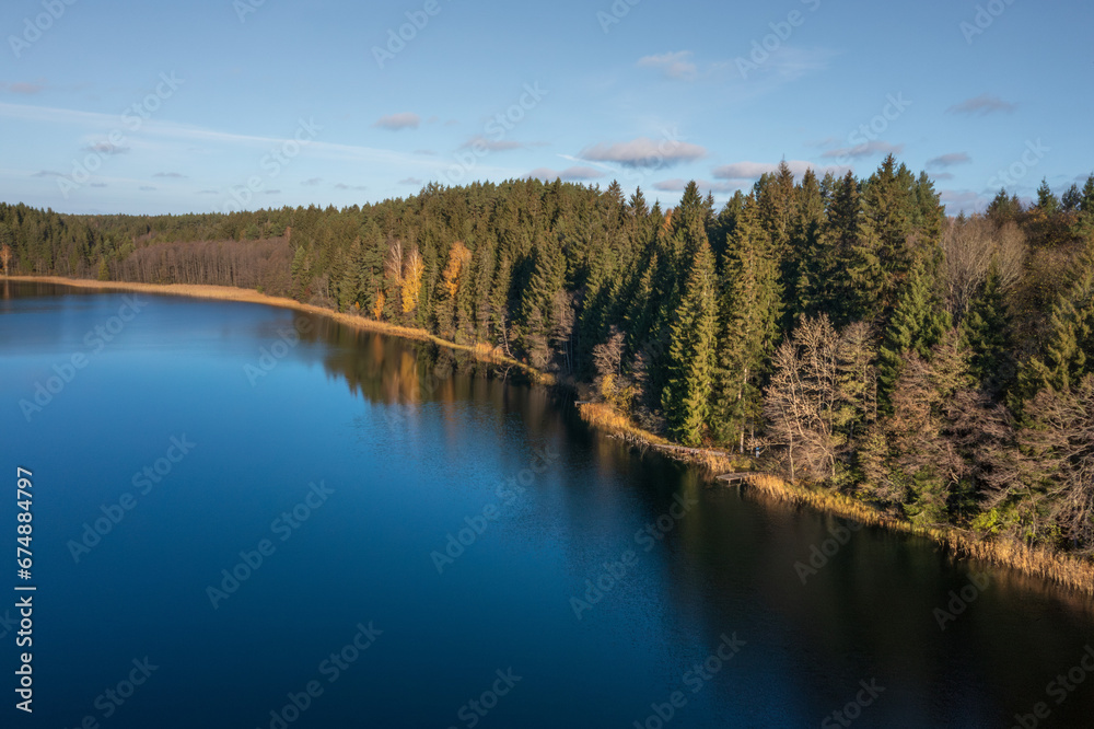 Aerial view on bank lake Bolduk in the autumn sunny morning. Woodland and sky are reflected on the water of lake. Small wooden piers on the right bank of the lake.