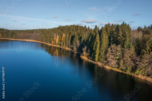  Aerial view on bank lake Bolduk in the autumn sunny morning. Woodland and sky are reflected on the water of lake. Small wooden piers on the right bank of the lake.