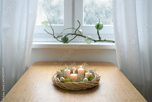 Homemade advent wreath with Christmas balls, moss and white candles, the second one is lit, on a table near the window on a snowy winter day, copy space, selected focus photo