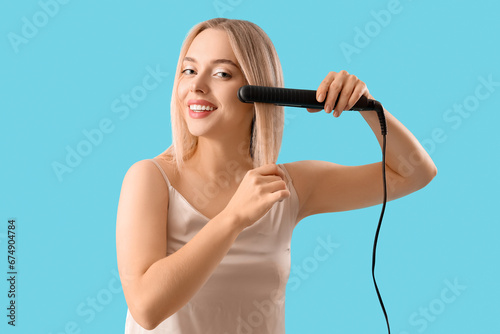 Young blonde woman straightening hair on blue background