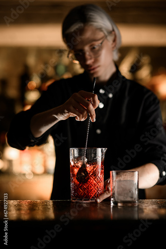 Female bartender mixes a cocktail in a mixing glass with ice, using a bar spoon