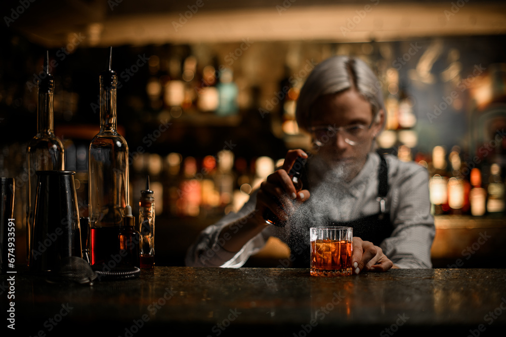 Female bartender squatting at the level of a glass sprays a ready-made cocktail in a glass