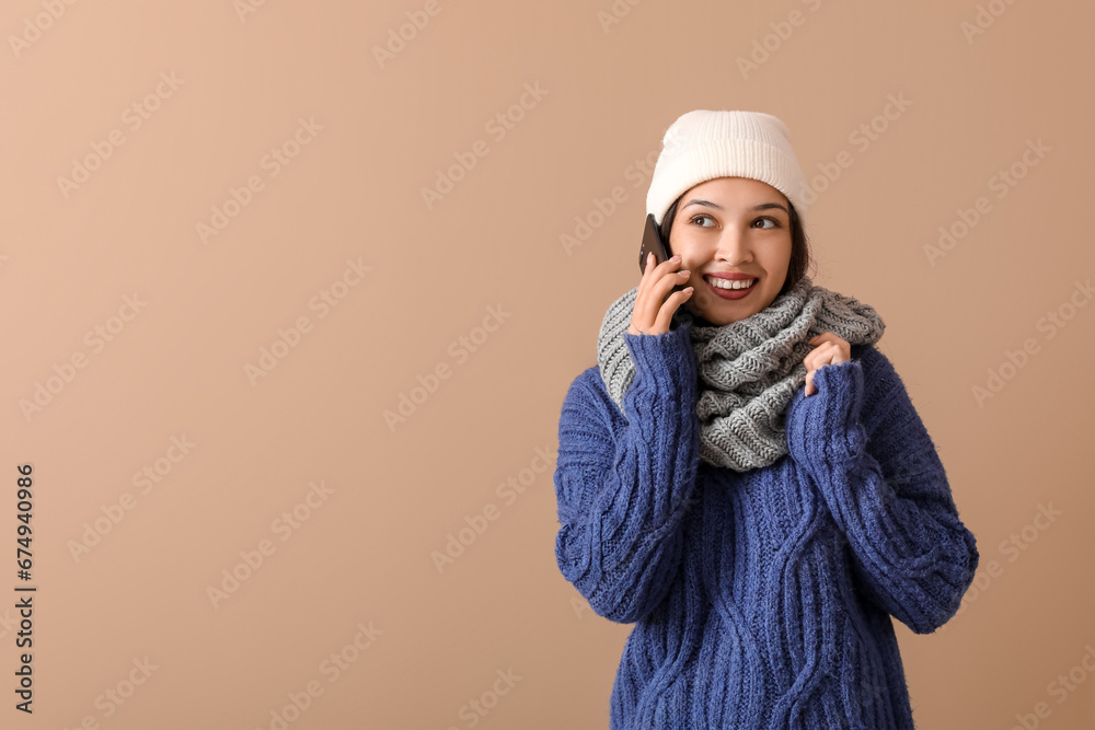 Beautiful young Asian woman in warm scarf talking by mobile phone on brown background