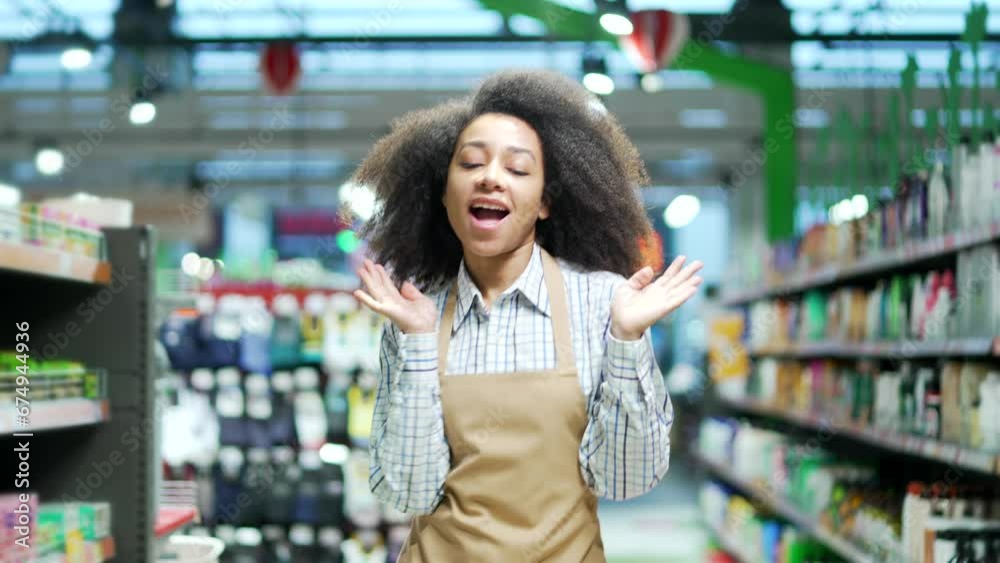 Joyful supermarket saleswoman worker dancing at marketplace. Happy funny black woman seller in apron rejoices at workplace between rows of shelves in market grocery store Cheerful glad employee 