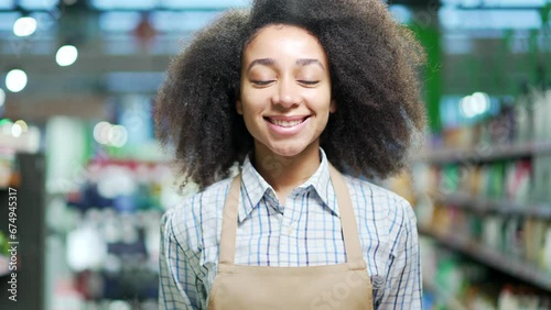 Portrait of a saleswoman female worker in a supermarket looking at camera, smiling. African american black woman employee clerk in apron in grocery store Happy Positive girl in market indoor Closeup photo