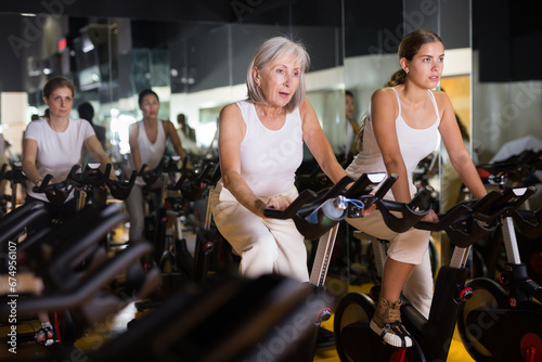 Young and mature women doing cardio workout at gym, training together on exercise bikes