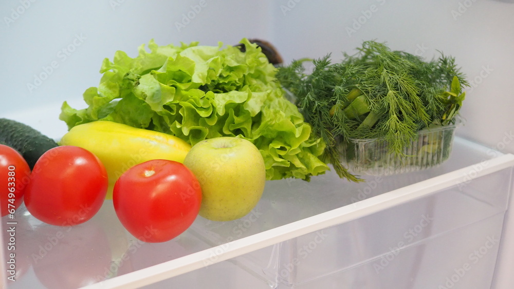 Woman hand taking, grabbing or picks up fresh fruits, vegetables and herbs out of open refrigerator shelf or fridge drawer full of fruits, vegetables, apple, tomatoes, cucumbers, lettuce. Healthy food