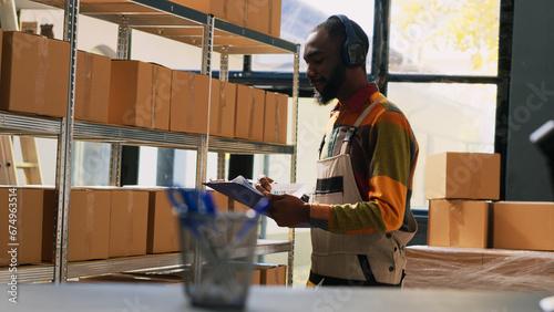 Male worker listening music in depot, working with merchandise stock in storage room. Warehouse manager checking inventory list on papers, planning quality control and distribution.