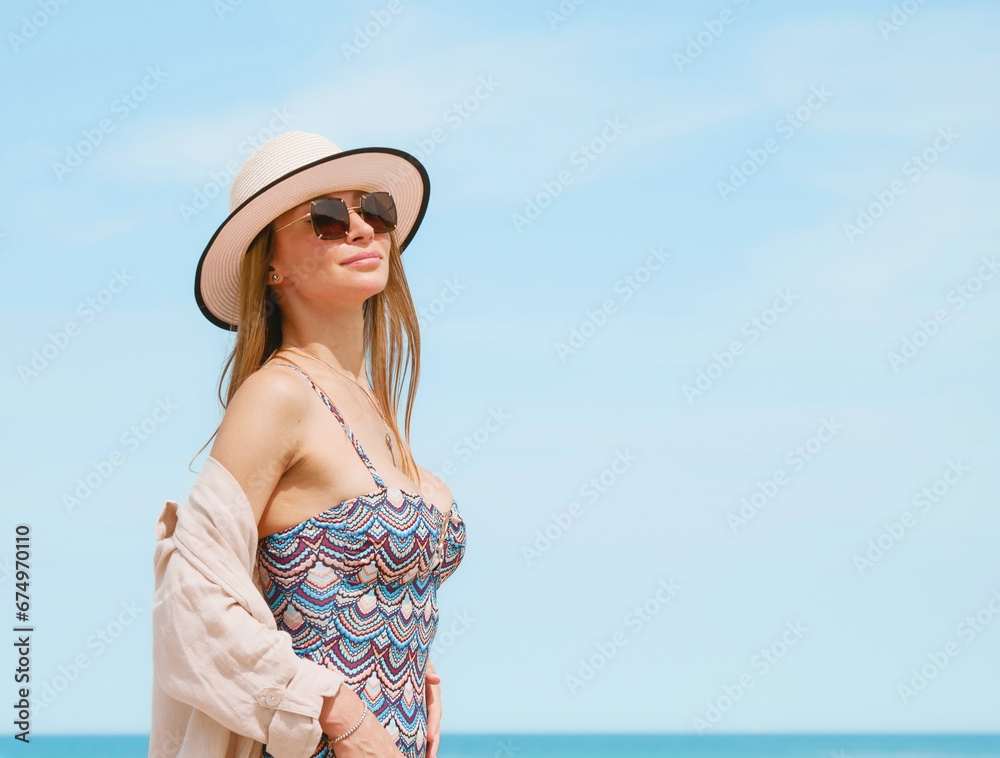 Young woman in a swimsuit, shirt, sunglasses and hat posing gracefully on a sandy beach against a blue sky