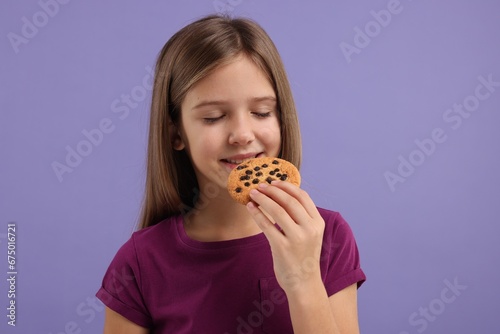 Cute girl eating chocolate chip cookie on purple background