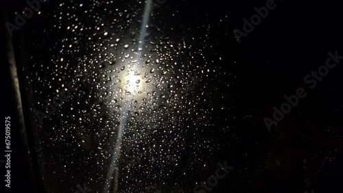 Car glass in raindrops. Rain in the night. Puddle. Streetlight. Rain drops Falling On puddle. Abstract bokeh lights of traffic in night time behind the wet window in slow motion