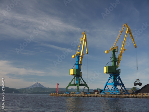 Industrial scene with Two harbor cranes with big lifting grabs loading coal on cargo terminal in Kamchatka harbor, Vilyuchinsky volcano in background photo