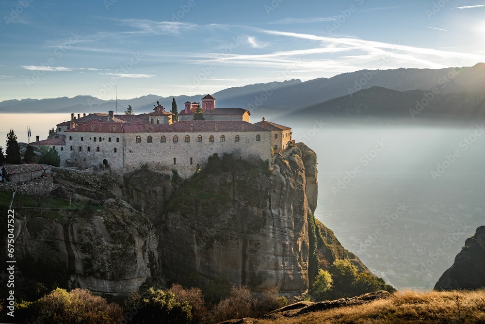 Historic monastery atop a mountain cliff, overlooking a tranquil lake in Meteora, Greece.