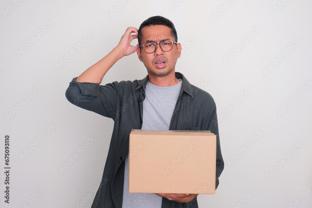 Adult Asian man showing confuse expression while holding a box of package Adult Asian man showing confuse expression while holding a box of package