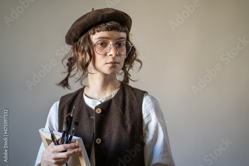 Serious illustrator in vintage clothes holding canvas, paint brushes, looking at camera. Young artist preparing for work, drawing, creating sketches. Profession of drawing tutor, illustrator, painter