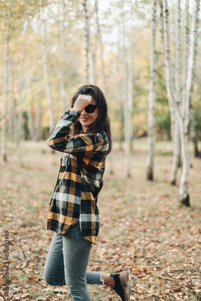 A beautiful girl in jeans and a plaid shirt walks in a birch grove. Portraits of a brunette girl in nature, in the autumn forest A beautiful girl in jeans and a plaid shirt walks in a birch grove. Portraits of a brunette girl in nature, in the autumn forest