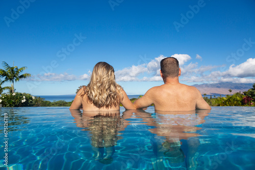 Rear view of handsome couple in an infinity pool looking out tropical island landscape in Hawaii. View of the Ocean on a beautiful island. Travel, happiness, luxury, summer holiday concept.