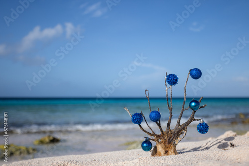 Fototapeta Naklejka Na Ścianę i Meble -  Dead corals decorated with blue Christmas balls standing on the sand beach with blue sea behind it 