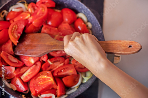girl stirring tomatoes with onions with a special wooden spatula for cooking in a frying pan in the kitchen. flatlay © Nataliia Makarovska