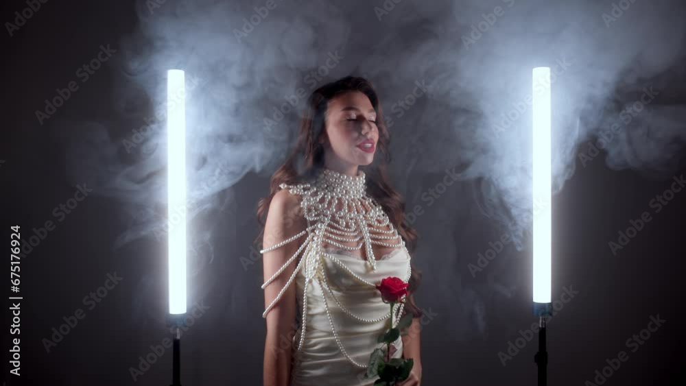 Beautiful young woman on a dark studio background in smoke with a red rose in her hands for Valentine's Day, women's empowerment or self-love. Beauty model with cosmetics or stylish clothes. Beautiful young woman on a dark studio background in smoke with a red rose in her hands for Valentine's Day, women's empowerment or self-love. Beauty model with cosmetics or stylish clothes.