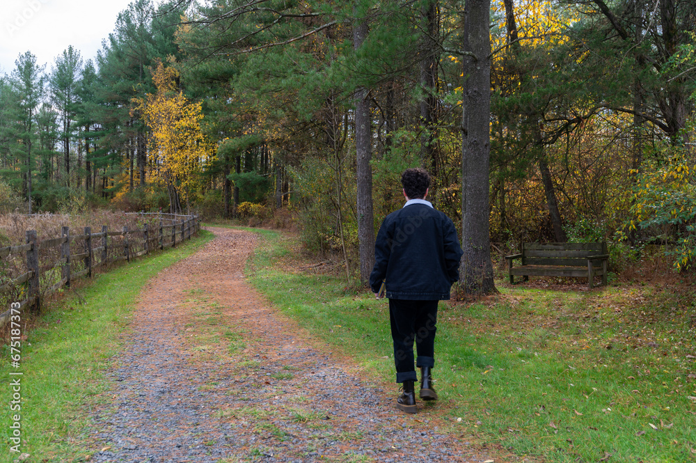 Young man in black walks away alone down country dirt road with autumn foliage on trees and split rail wooden fence. With copy space