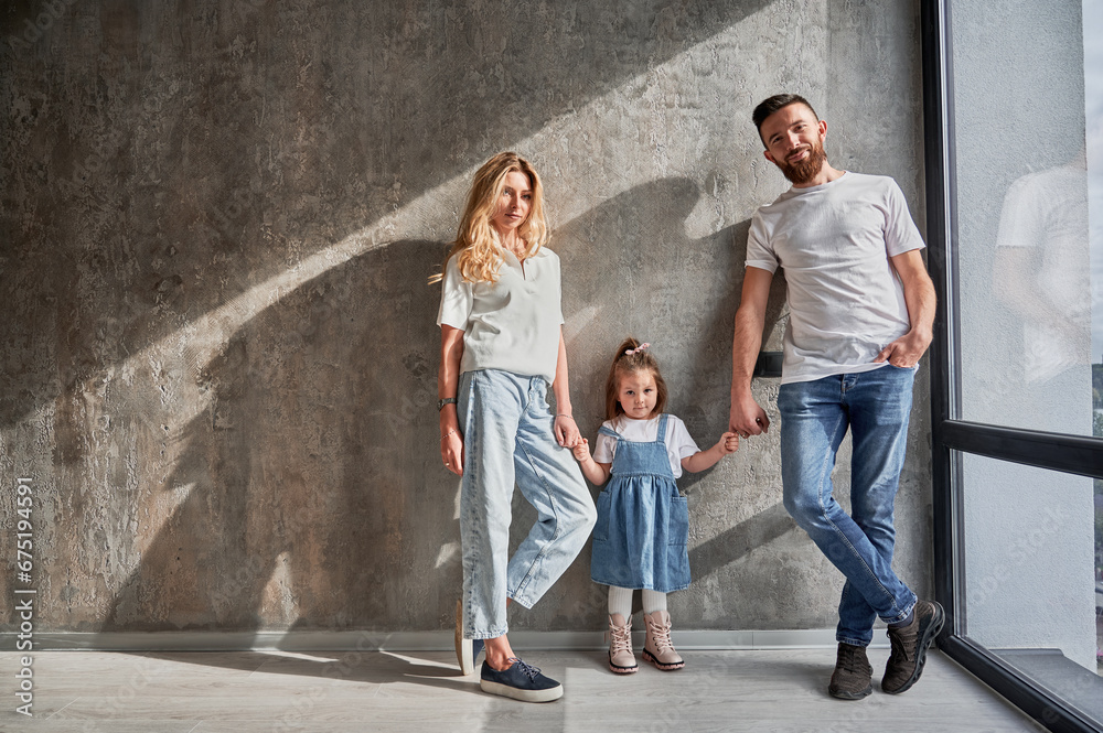 Portrait of happy parents and child standing by the window against the wall in apartment. Man and woman holding hands of adorable daughter in new home with sunlight shadow on the wall.