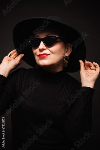 Trendy mature woman with red lips posing in hat, sunglasses and turtleneck isolated on black 