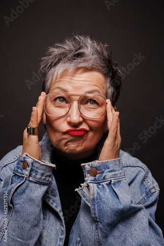 Trendy mature woman in eyeglasses and denim jacket grimacing isolated on black 