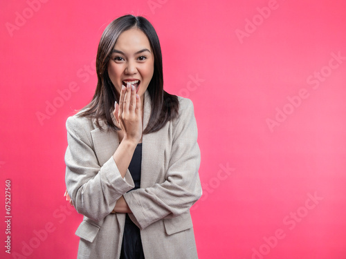 A portrait of an Indonesian Asian woman wearing a cream-colored blazer, posing in surprise and covering her mouth. Isolated against a magenta background.