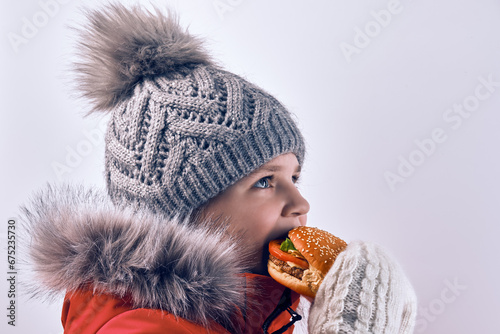 A 10-11 year old girl eats a hamburger in winter clothes, against a light background in the studio. photo