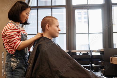 Side view portrait of smiling young woman with buzzcut sitting in salon chair with hairstylist working