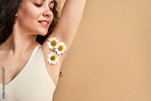 Femininity concept. Close up of curly haired woman in comfy white bra posing over beige background with chamomiles on smooth armpit. Caucasian young lady satisfied with effect after epilation.