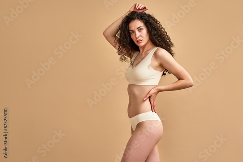 Self acceptance. Tender young woman dressed in lingerie touching head and slender waist with hands while looking confidently at camera. Isolated over beige studio background with copy space.