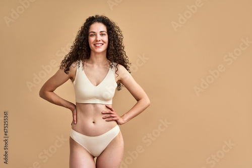 Feminine vitality. Happy young woman in bikini with hands on slim waist posing over beige studio background. Beautiful caucasian lady with wavy dark hair smiling and looking at camera.