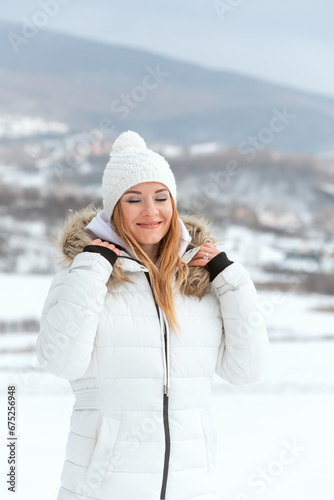 Portrait of attractive young woman in winter in mountains. Girl wears white down jacket and knitted hat and smiles. Vertical frame.