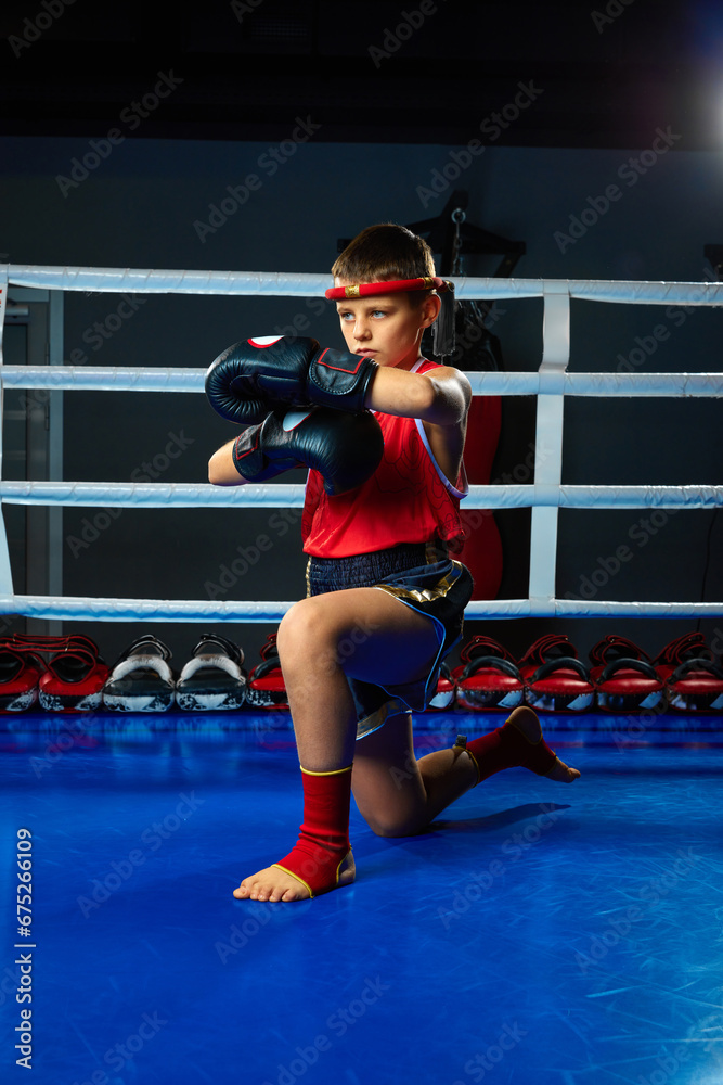 Serious boy, kickbox pupil, martial arts sportsman wearing uniform stands on knee and posing before fights.