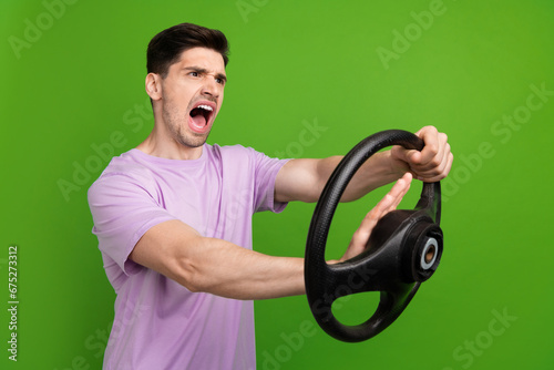 Photo of crazy man with brunet hair dressed purple t-shirt hold steering wheel scream in empty space isolated on green color background