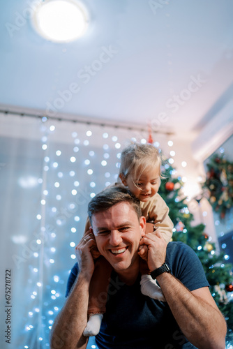 Laughing dad with a little girl on his shoulders leaned forward sitting near the Christmas tree