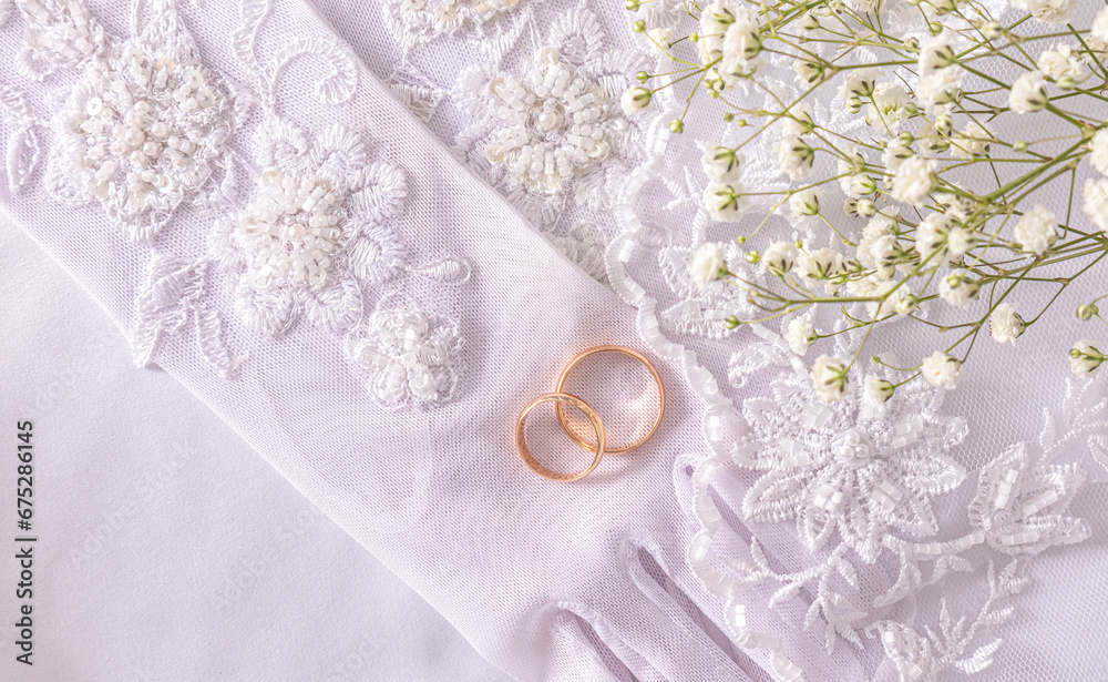 Two gold wedding rings lie on the bride's chic beaded gloves embroidered with beads and sequins. Top view. Satin white background.