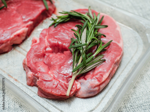 Juicy uncooked lamb leg steak with green fresh rosemary on plastic tray. Uncooked high quality meat product with rich taste and vivid color.