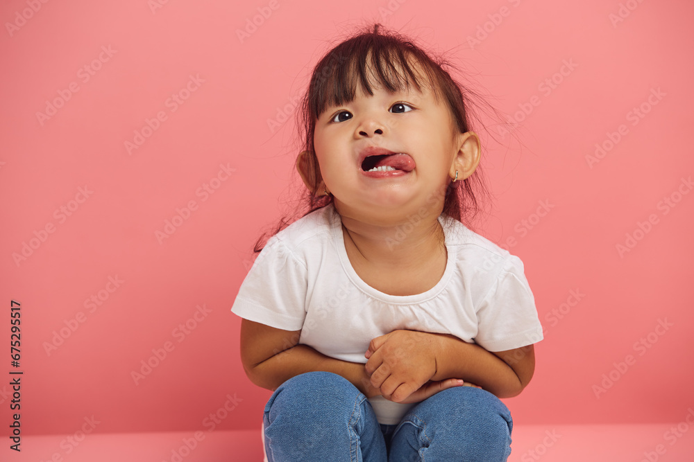 Funny little child girl capriciously shows his tongue, sits on the floor on a pink isolated background, close up portrait. Funny little child girl capriciously shows his tongue, sits on the floor on a pink isolated background, close up portrait.