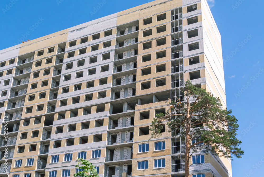 A pine tree grows in front of the house under construction. The high-rise residential building is lined with decorative bricks. The frame of a house under construction without window frames.