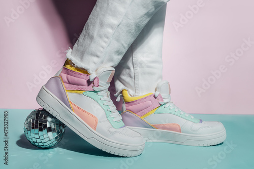 Close up photo of legs wearing retro style high-top multicolor sport sneakers shoes with disco ball on blue and pink background. Minimalism party, vintage retro style of 80s - 90s vibes.
