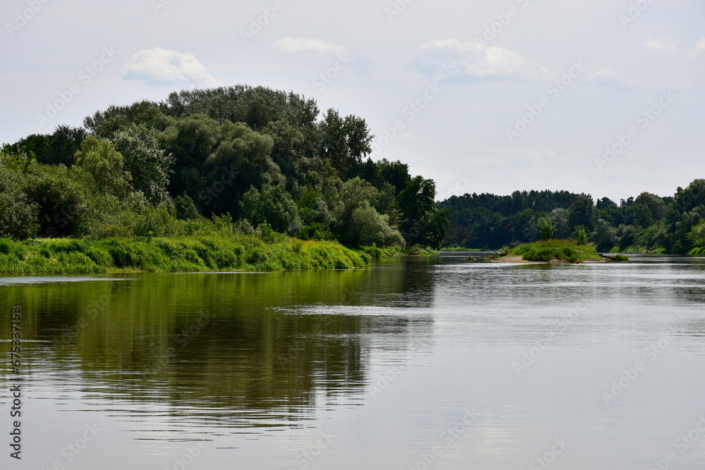 View of a massive lake, river, or pond surrounded from all sides with reeds, moss, forests, moors, and other flora spotted on a cloudy yet warm summer day next to some holiday resort located in Poland