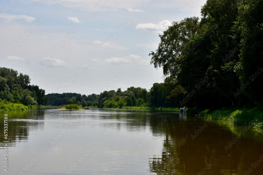 Fototapeta premium View of a massive lake, river, or pond surrounded from all sides with reeds, moss, forests, moors, and other flora spotted on a cloudy yet warm summer day next to some holiday resort located in Poland