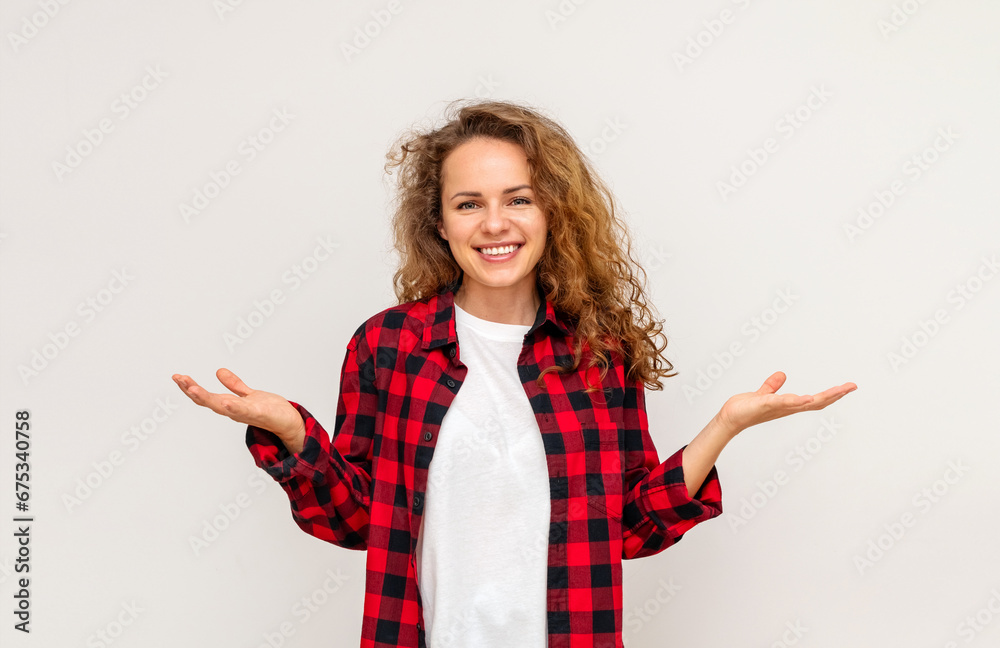 Portrait of a smiling young curly woman wearing a checkered red shirt standing with outstretched arms, palms up in front of white backdrop in studio. Portrait of a smiling young curly woman wearing a checkered red shirt standing with outstretched arms, palms up in front of white backdrop in studio.
